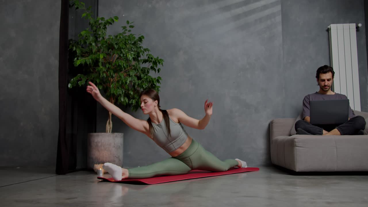 A confident brunette girl in a gray top and green sweatpants is doing stretching and sitting in the splits on a red sports mat near her her boyfriend who is doing work using a gray laptop at home in a modern apartment