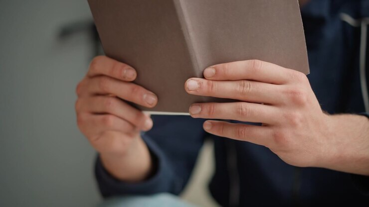 Detailní záběr rukou držících hnědou knihu s měkkým osvětlením a neutrálním pozadím. Osoba má na sobě tmavé oblečení, zaměřující se na interakci mezi prsty a předmětem. Pozadí tvoří obyčejná šedá zeď.