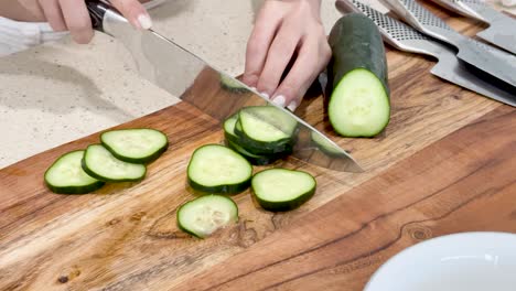 a person slices a cucumber into even rounds on a wooden cutting board in a bright kitchen, using a large chef’s knife and natural lighting