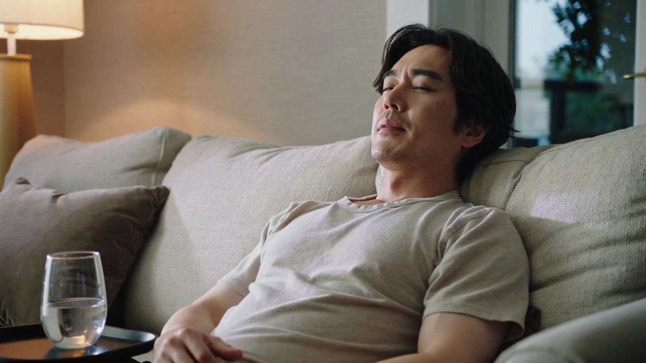 Wincing, Asian man in light T-shirt rubbing temple, leaning on sofa at home with water glass