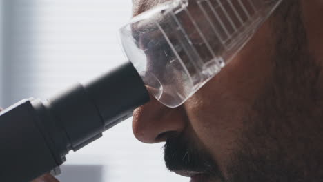 close up of male scientist in goggles looking through microscope eyepiece