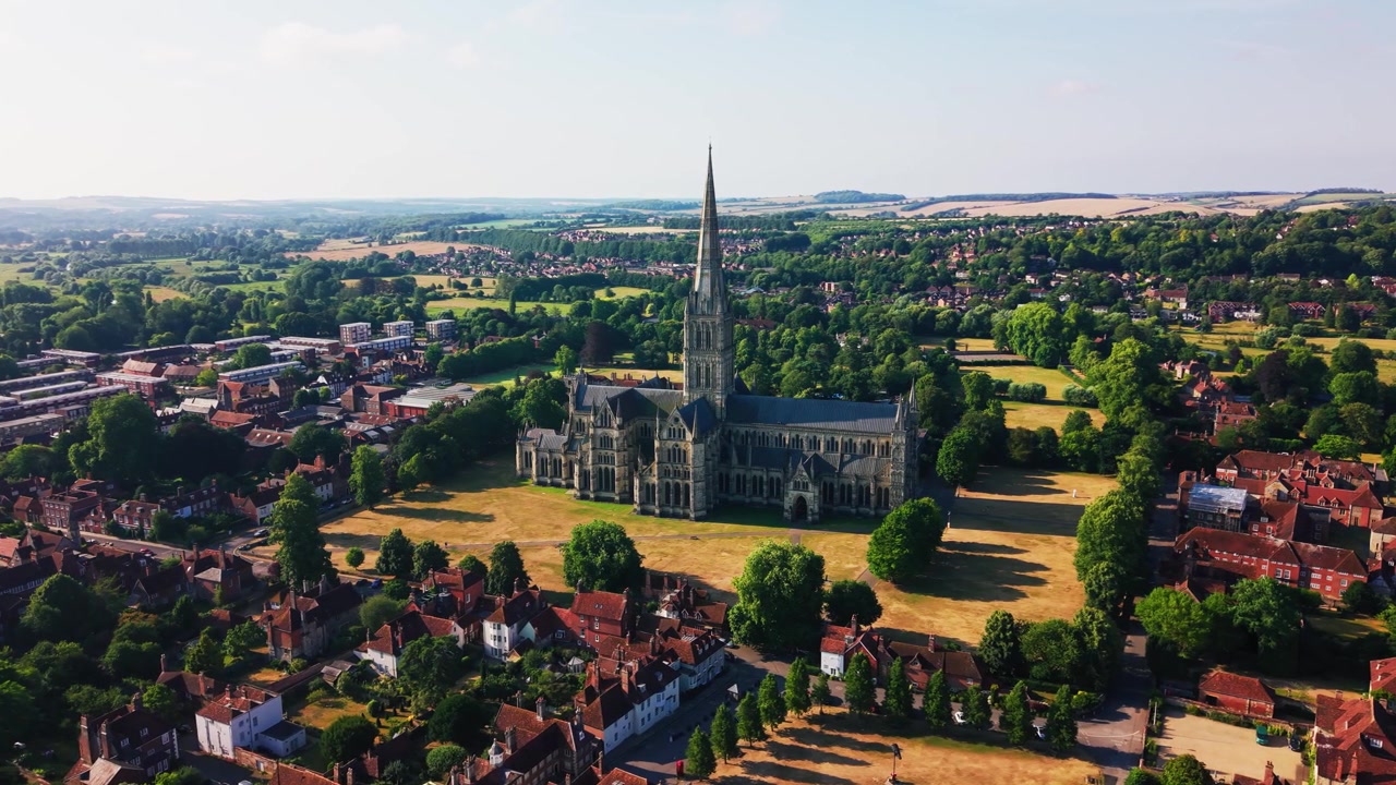 Szeroki widok z drona na katedrę w Salisbury i jej średniowieczną iglicę otoczoną historycznymi dachami, zielonymi terenami i wiejskimi krajobrazami Wiltshire w słoneczny letni dzień