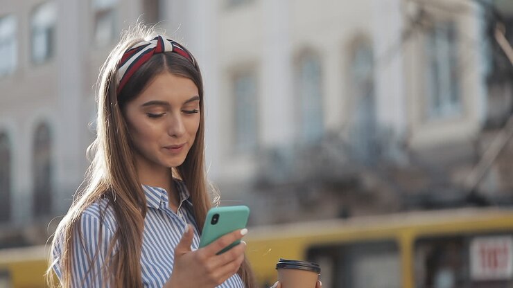 Giovane donna felice che scrive messaggi sullo smartphone e tiene un caffè in città
