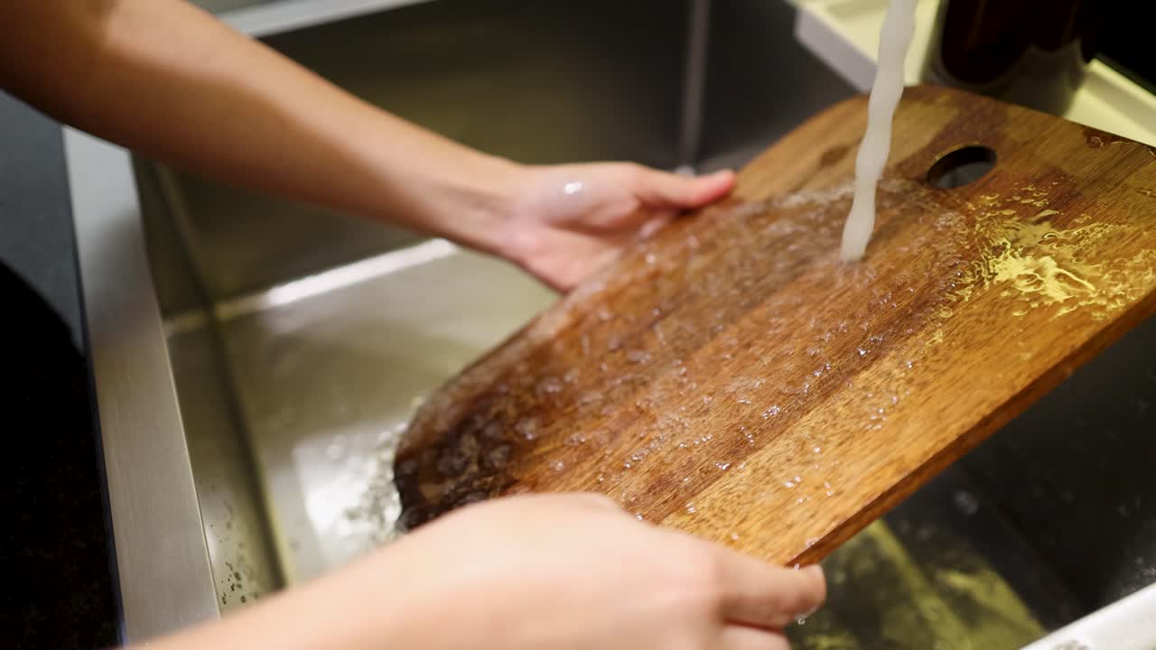 Close-up shot of hands rinsing a dark wooden cutting board under a kitchen faucet stream