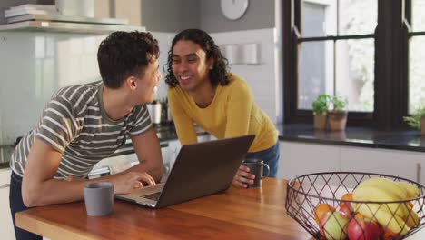 feliz pareja de hombres diversos bebiendo café y usando computadora portátil en la cocina