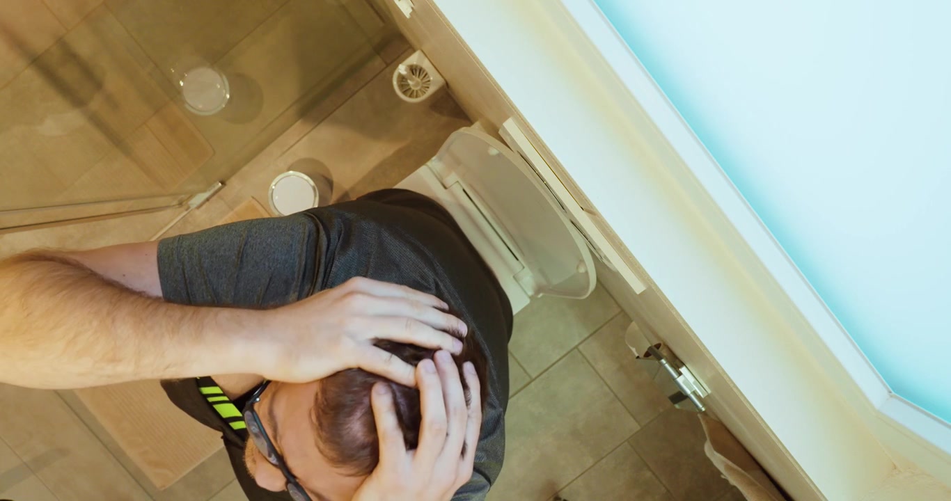 Top view of a man sitting on a toilet while holding his stomach in pain. Concept of abdominal cramps, digestive problems, illness, and bathroom emergency.