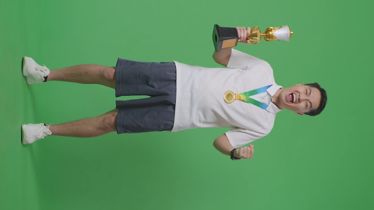 황금 메달과 트로피를 가진 아시아 남성의 몸이 가득 차 있으며, 골을 외치며 스튜디오의 녹색 스크린 배경에서 첫 번째 우승자로 우승을 축하하고 있습니다.