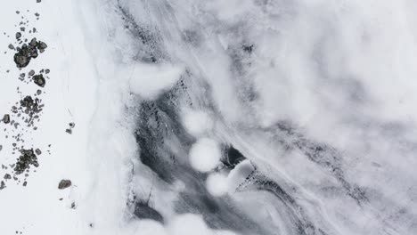 aerial drone footage flying directly above beautiful ice formations in a frozen mountain loch with scattered rocks and boulders frozen in the ice