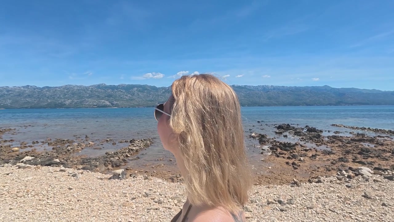 Close-up of a woman standing on a sunny beach while enjoying the fresh sea breeze. Relaxing coastal atmosphere with clear water, summer sunshine, and vacation vibes.