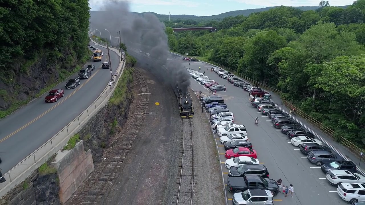 Una locomotiva a vapore per treni passeggeri emette fumo mentre passa lungo una strada. Auto sono parcheggiate nelle vicinanze e il paesaggio lussureggiante crea uno sfondo naturale in una calda giornata estiva.