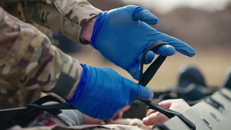 close up a confident male doctor in a camouflage uniform with blue medical gloves unties the straps on a stretcher for unconscious wounded soldiers during combat operations in the steppe