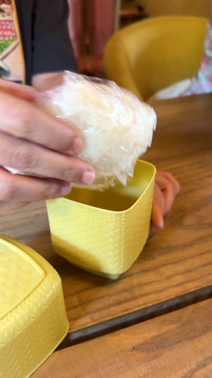 Mão remove arroz pegajoso embalado em plástico de uma cesta amarela em uma mesa de madeira, iluminação natural interna.