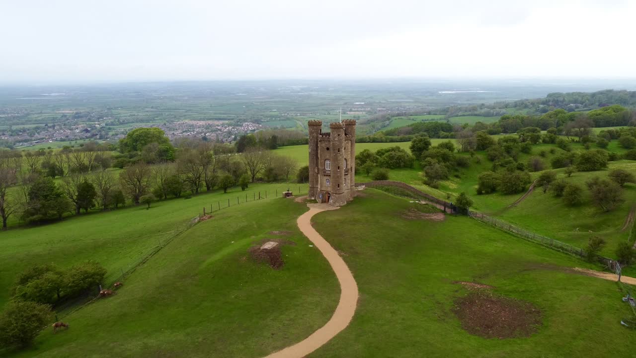 Aerial view of Broadway Tower in the Cotswolds, England, standing on a grassy hilltop surrounded by scenic countryside and pathways, showcasing historic charm and panoramic rural landscape