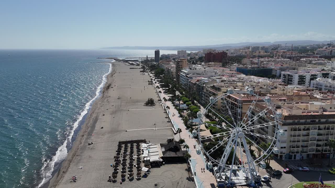 Drone shot of long sandy beach beside coastal promenade with Ferris wheel, umbrellas, and city buildings stretching into distance under bright sky, blending urban life with seaside relaxation.