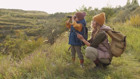 Young-Female-Backpacker-Squatting-By-Her-Adorable-Little-Son-With-Binoculars-During-Weekend-Trip-In-Natural-Environment