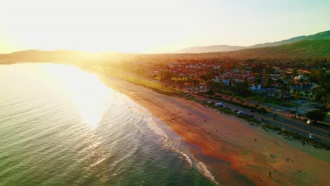 Aerial view of Santa Barbara, California at golden hour, showcasing the coastline, palm-lined road, and nearby homes bathed in warm sunlight.