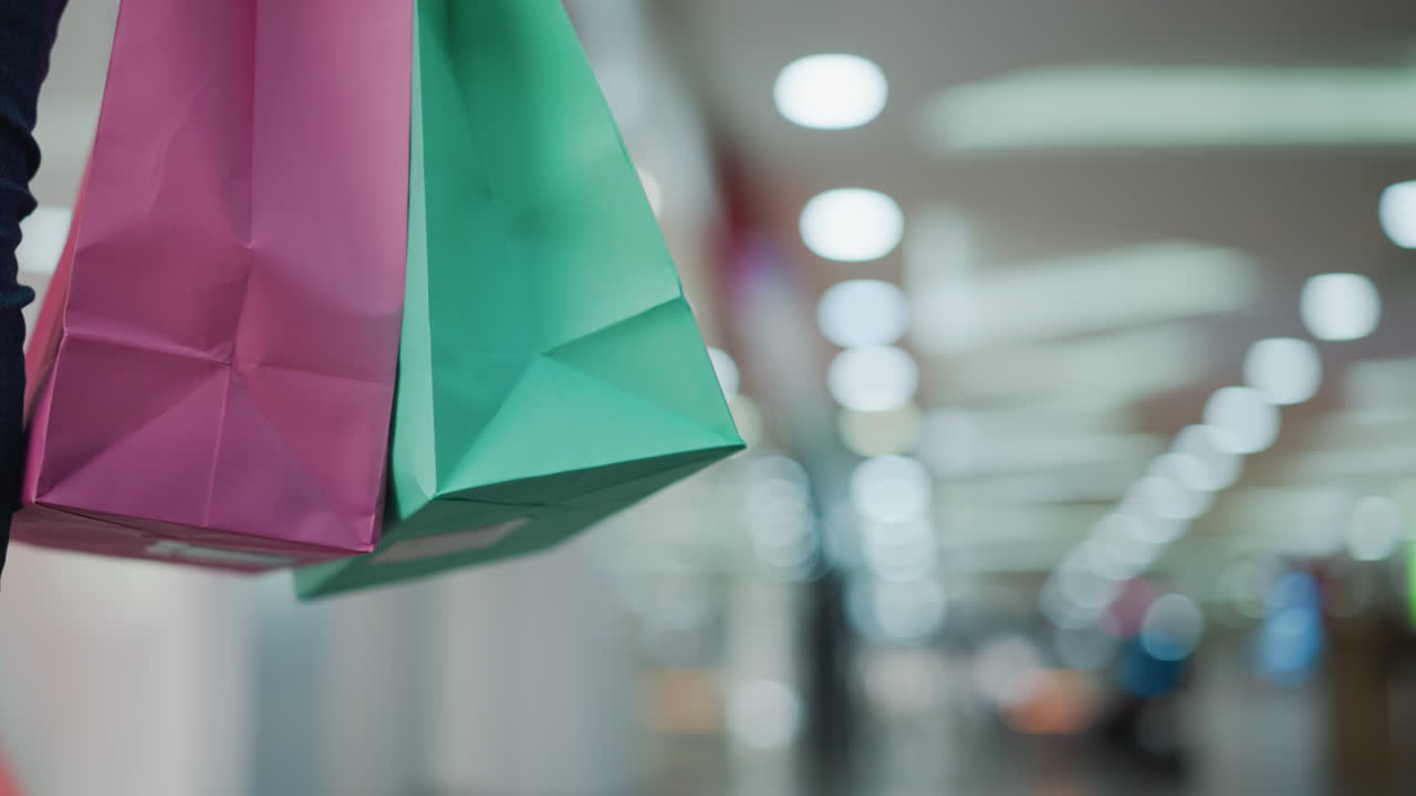 primer plano de bolsas de compras rosas y verdes llevadas por una persona en vaqueros caminando a través de un centro comercial brillantemente iluminado con luces borrosas y colores vibrantes en el fondo