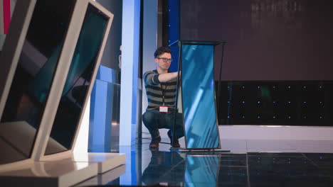 man crouches down adjusting digital display screen on studio floor, preparing equipment for video production, reflective blue tiles, with background lights and colorful studio set in view