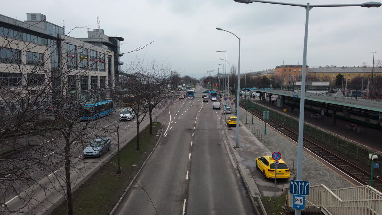 Weitwinkel-Luftaufnahme einer belebten Straße mit gelben Taxis und blauen Bussen am Örs vezér tere. Simulierte Drohnenperspektive des Budapester Nahverkehrs in realistischer 2026er-Ästhetik.