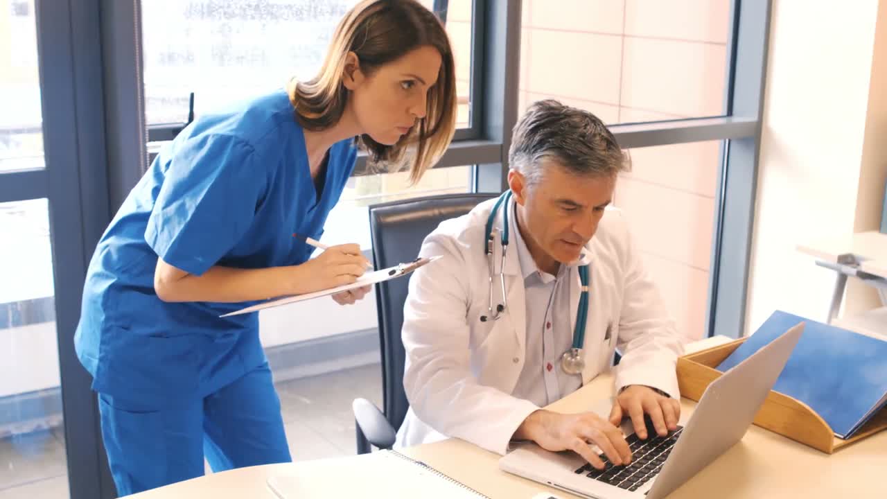 médico usando computadora portátil mientras la enfermera escribe en el clipboard