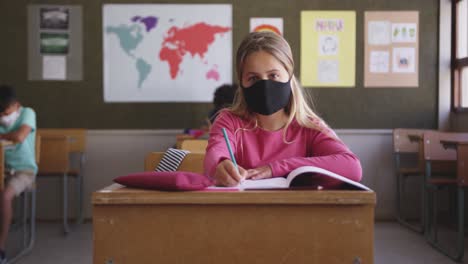 Mädchen-Mit-Gesichtsmaske-Schreibt,-Während-Sie-In-Der-Schule-Auf-Ihrem-Schreibtisch-Sitzt