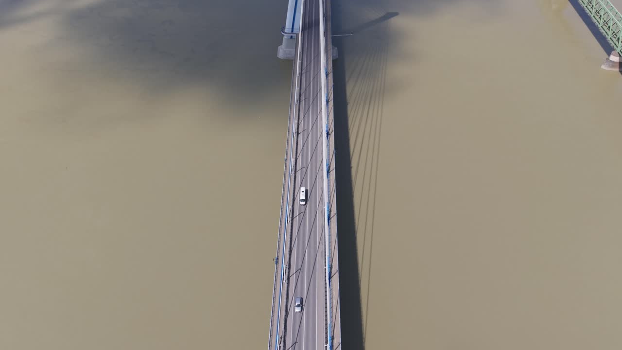 A vibrant pink truck and several cars cross a modern cable-stayed bridge spanning a muddy river. The bridge's sleek structure and surrounding farmland are visible from an aerial perspective