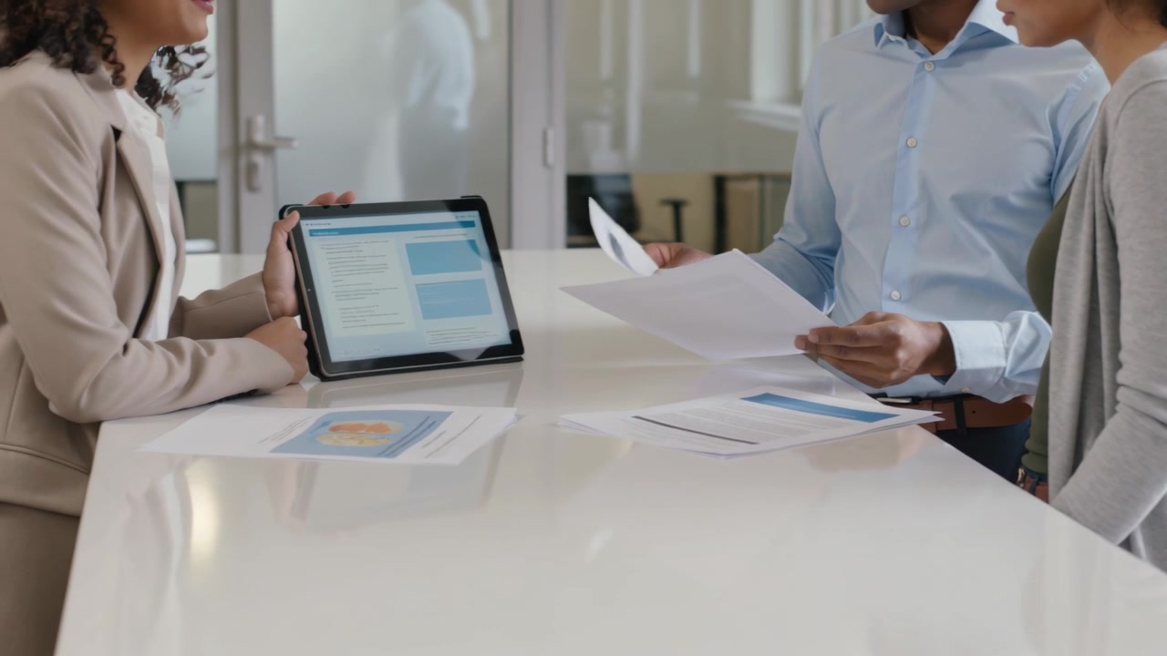 Propping tablet, left woman presenting while two colleagues flipping reports at office table, suits
