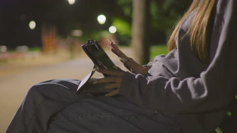 close-up of young girl sitting on park bench at night, fully focused on tablet under soft, serene park lighting, blurred background reveals people strolling