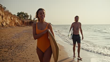 una chica rubia feliz y confiada en un traje de baño naranja camina a lo largo de la costa rocosa y arenosa y lleva una tabla de surf detrás de ella, su novio rubio en pantalones negros está caminando a lo largo que también está llevando una tablas de surf cerca del mar en la orilla por la mañana