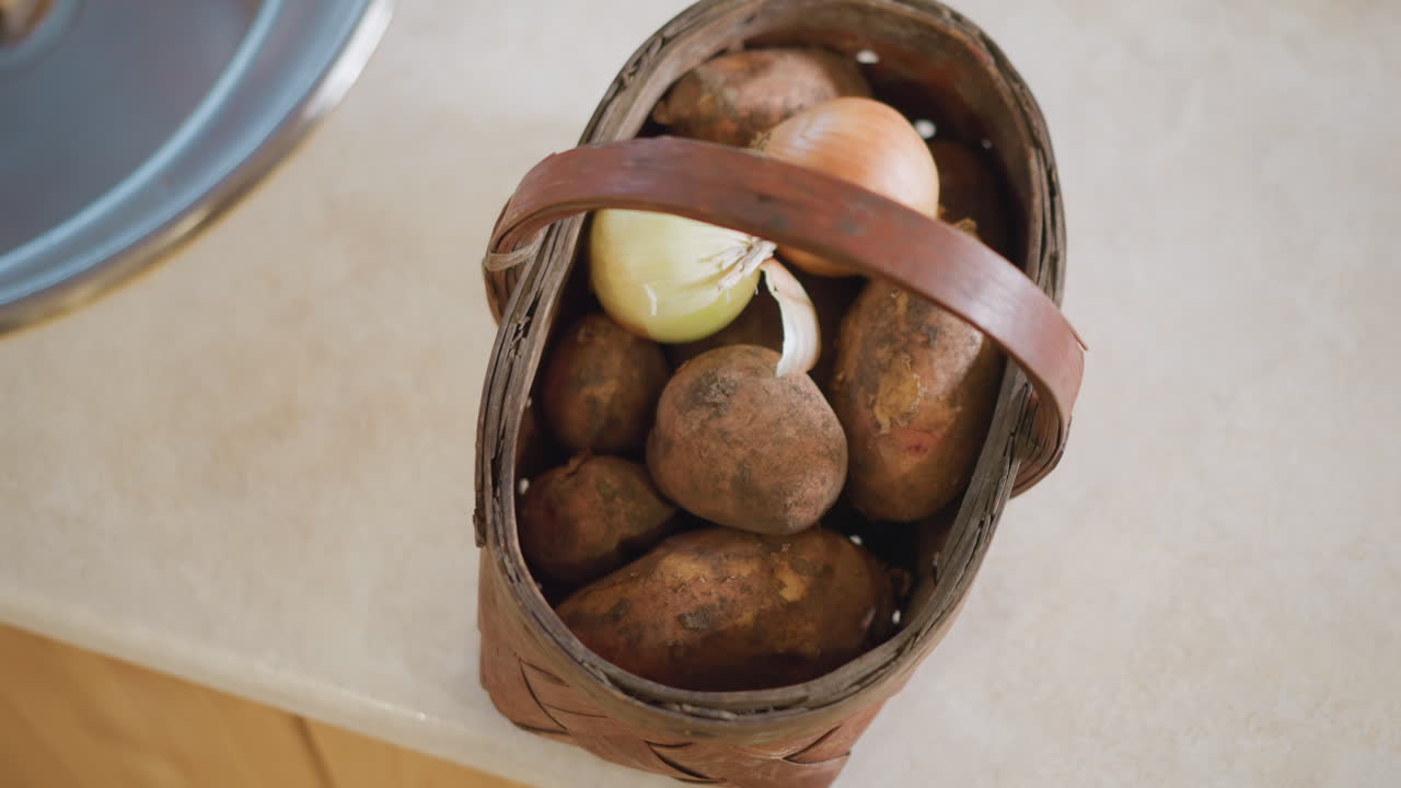 Vista superior de cesta de batatas na bancada, alça de couro e cebola visíveis, borda da pia na extremidade, colheita prática de cozinheira doméstica preparada para ferver ou assar com configuração de cozinha simples