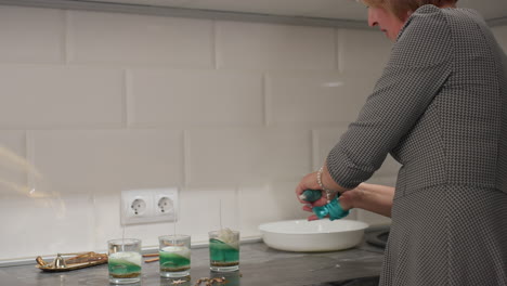 female artist in fitted patterned dress squeezes leather-like material with knot detail on countertop near three green candle jars, white ceramic bowl, and golden tray, focused on creative process
