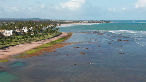 Vista-Aérea-De-La-Playa,-Olas-Y-Una-Gran-Zona-Verde-Con-Palmeras,-Una-Persona-Practicando-Kitesurf-Y-La-Ciudad-Al-Fondo,-Guarajuba,-Bahía,-Brasil