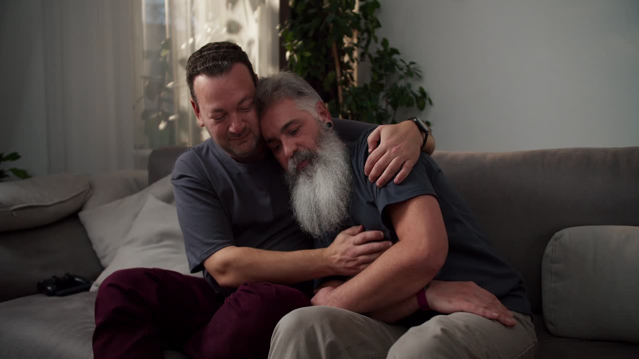 A middle-aged brunette man with stubble in a gray T-shirt hugs his sad boyfriend a man with gray hair color and a lush gray beard while sitting on a sofa in a modern apartment in the evening. Showing love and care in difficult times to a couple of middle-aged LGBT men in a modern apartment