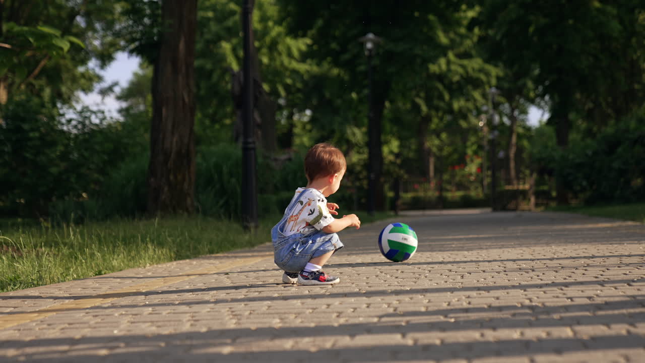 Little boy squats near the ball and pushes it to roll. Kid stands up and walks opposite side by the paved road.