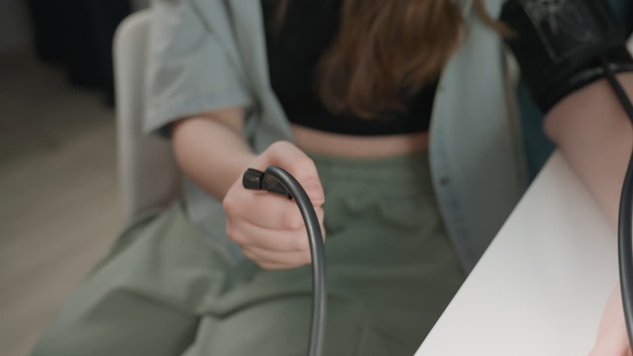 Young caucasian woman preparing blood pressure cuff on arm, sitting at white table with casual green trousers, holding tubing with tentative grip, natural soft light, focused expression during athome