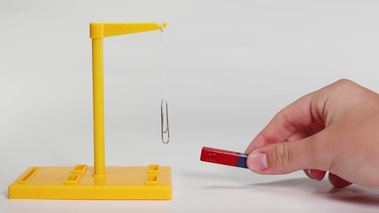 A hand manipulates a magnet near a suspended paperclip, demonstrating magnetic forces in a controlled environment with clear lighting