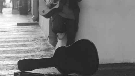 Vista-En-Blanco-Y-Negro-De-Una-Caja-De-Guitarra-En-El-Suelo-Y-Un-Hombre-Con-Abrigo-Cantando-Y-Tocando-La-Guitarra-Apoyado-En-La-Pared-De-La-Calle