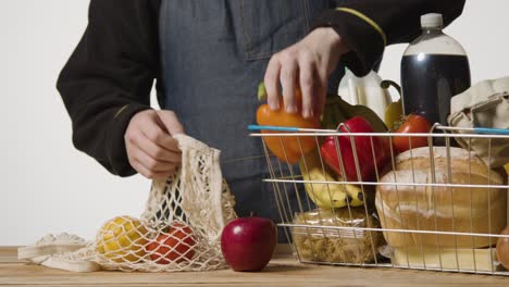 studio shot of shop worker packing basic food items from supermarket wire shopping basket into reusable bag