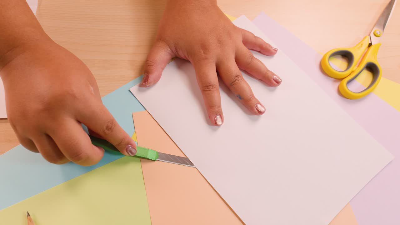 Person uses utility knife to cut and fold colored paper on tidy, well-lit workspace
