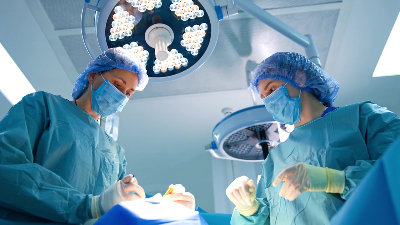 To kvindelige medicinere arbejder under en operation. Lav vinkel på lægerne, der påbegynder en operation. Runde lamper i baggrunden.