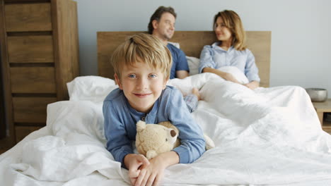 cute small boy lying with a teddy bear and smiling at camera, his parents are lying under the blanket on the bed behind him