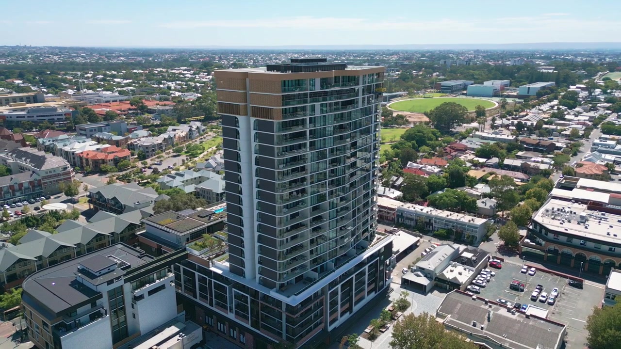 Ripresa orbitale attorno a una moderna torre residenziale a più piani con facciata in vetro che rivela un'espansione suburbana a bassa densità, un ovale sportivo e strade alberate in tutte le direzioni. Subiaco, Perth, Australia