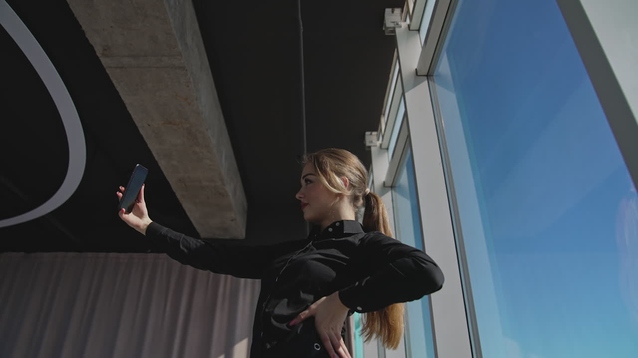 Successful confident millennial woman in black shirt smiling looking at camera, posing and making video. Female office worker making selfie in front of a big office window. View from below.