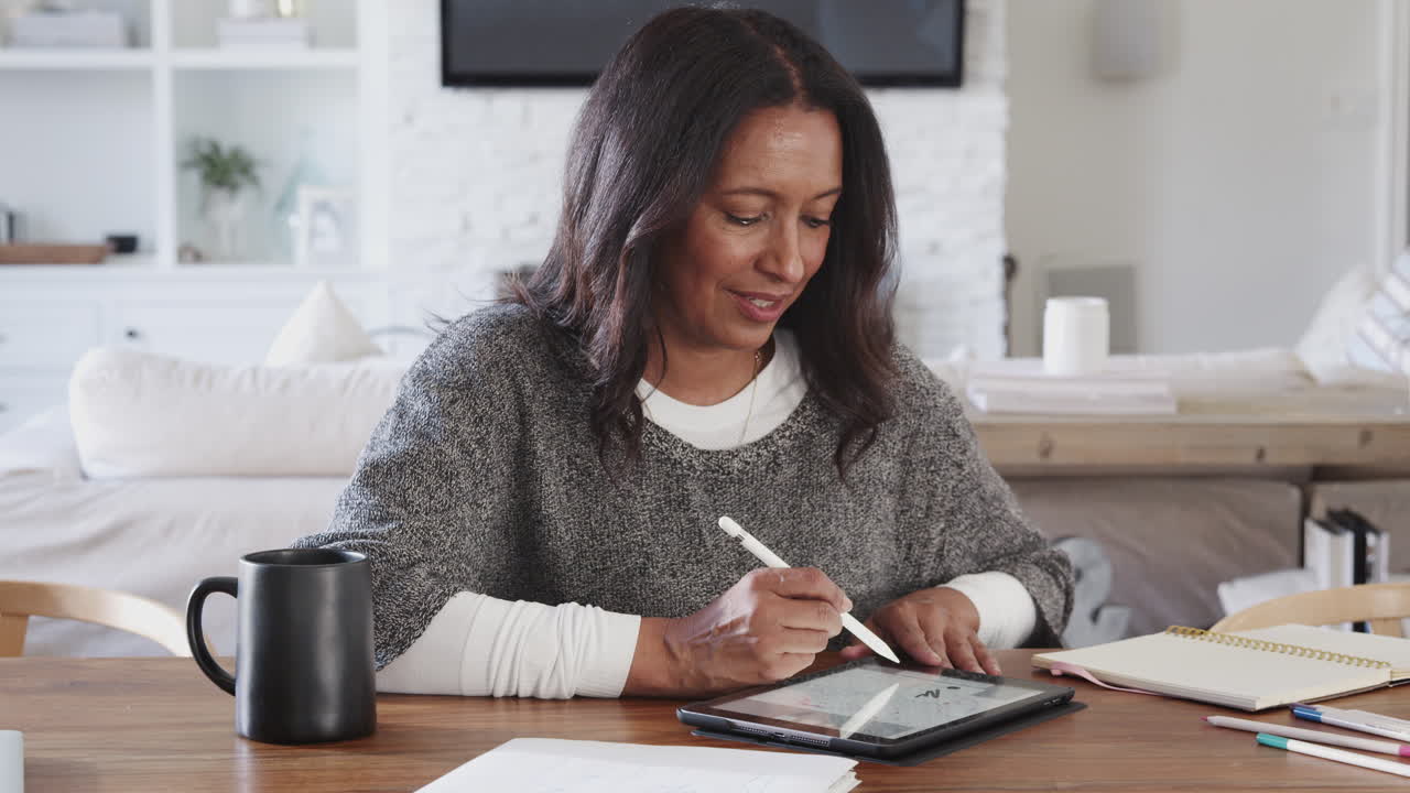 Middle aged woman sitting at a table using a tablet computer and stylus, front view, zoom out