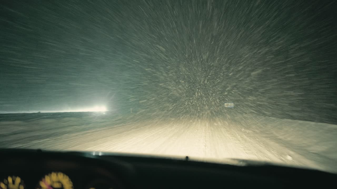 Ponto de vista da direção noturna filmado de dentro de um carro durante uma tempestade de neve em uma rodovia. Baixa visibilidade, estrada coberta de neve e faróis de veículos que passam mostram as duras condições de viagem no inverno.