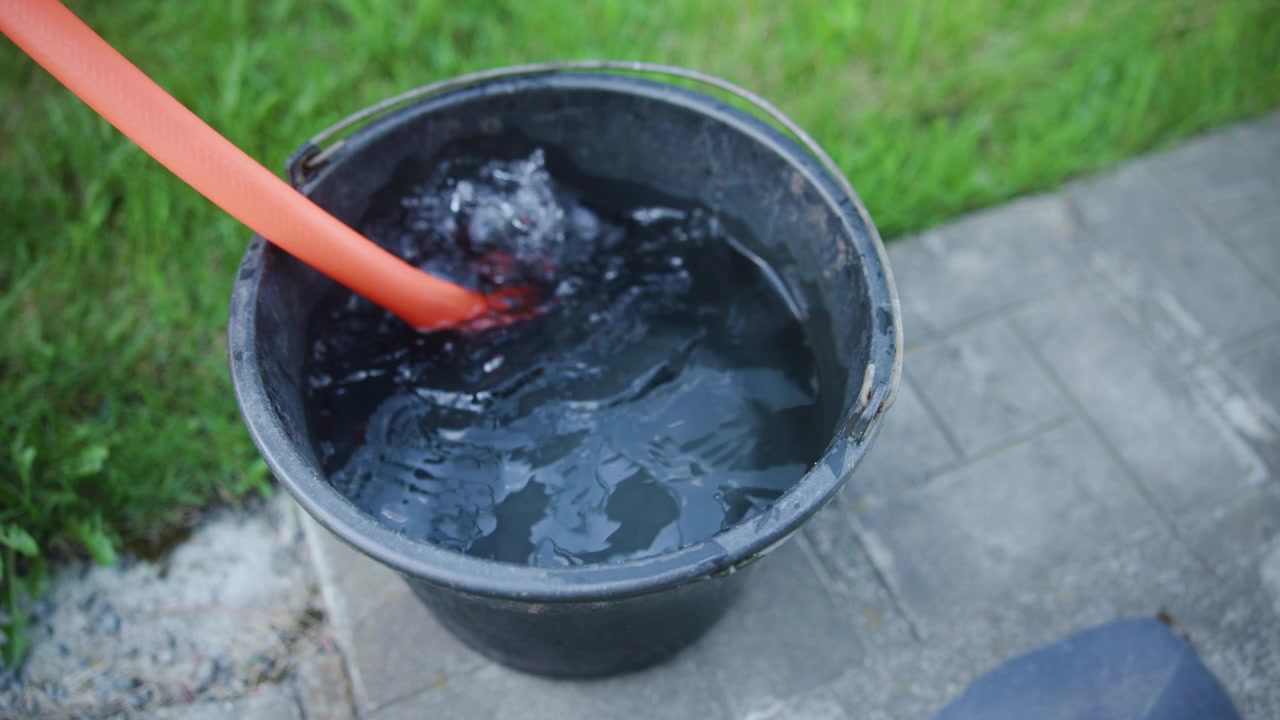 Ein orangefarbener Schlauch füllt einen schwarzen Plastikeimer auf einem Betonweg nahe grünem Gras, von oben gesehen bei sanftem Tageslicht mit wellenförmigen Reflexionen auf der Oberfläche.
