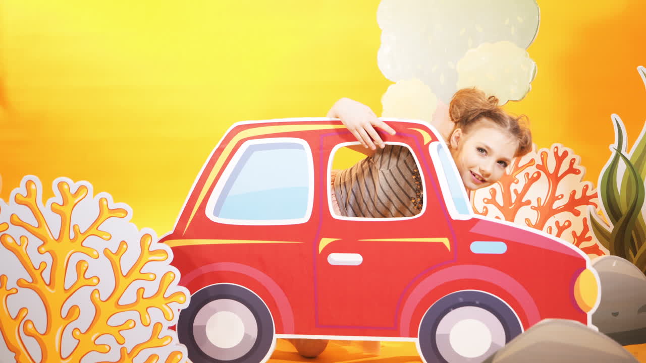 Funny little girl on a bright yellow wall background. A beautiful child is having fun with a big toy car.