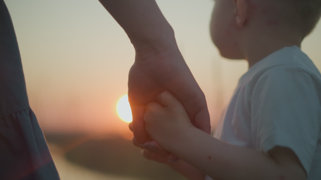 tender primer plano de una madre sosteniendo la mano de su hijo pequeño, que está vestido de blanco, mientras están juntos viendo una serena puesta de sol. el cálido resplandor del sol poniente se refleja en el tranquilo lago