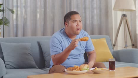 a fat asian man eating fast food having fried chicken while watching tv on a sofa in the living room at home