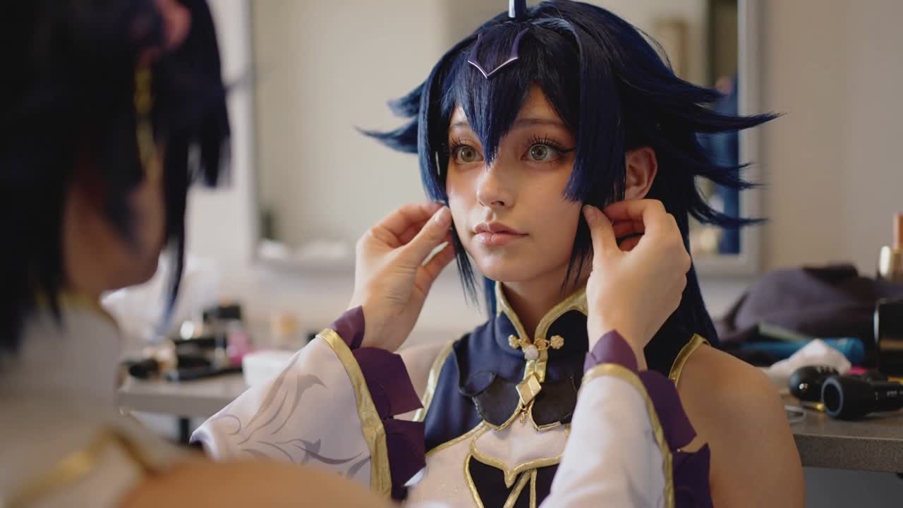 Người phụ tá đang với tay chỉnh tóc giả màu xanh và phụ kiện trên cosplayer trước gương, với chiếc cổ áo cầu kỳ để chụp ảnh.